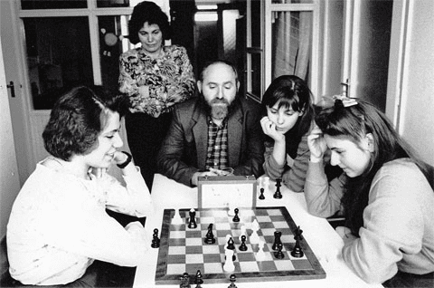 wokół szachów siedzą 3 siostry z tatą, za nimi stoi kobieta - siostry Polgar 